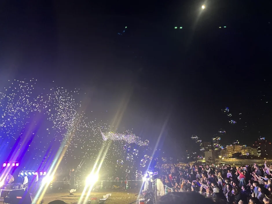 夜の屋外イベントで、多くの観客がステージの明かりと舞い上がるシャボン玉を楽しんでいる様子。空には月が見え、賑やかな雰囲気が伝わる。