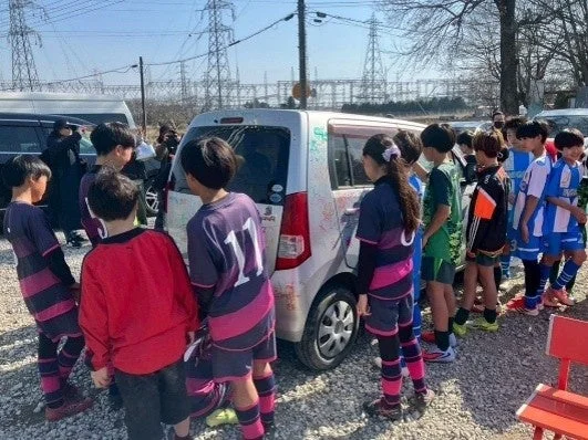 サッカーのユニフォームを着た子供たちが、屋外で銀色の車の周りに集まっています。