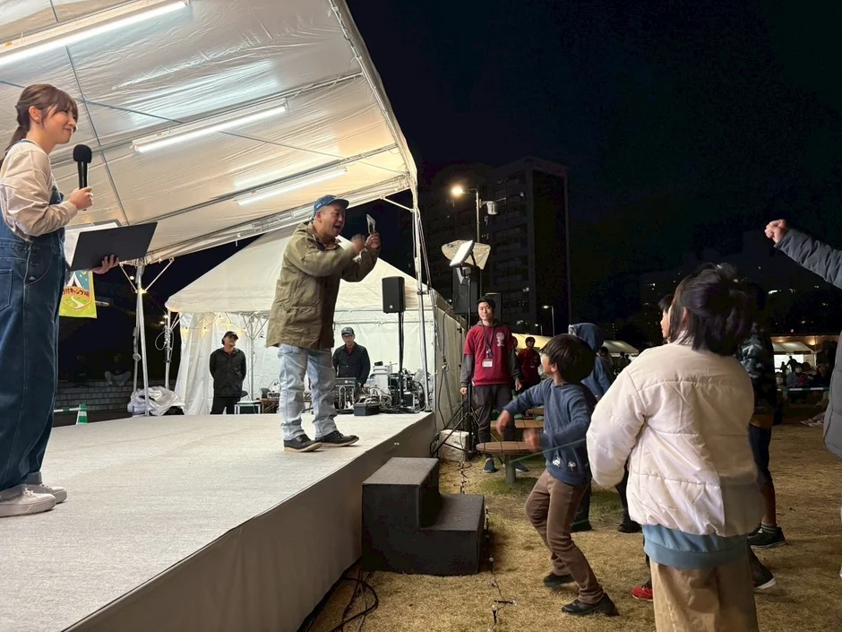 夜の屋外イベント会場で、テント下のステージに立つ女性司会者と男性パフォーマーを、子供を含む観客が見守っています。賑やかな雰囲気の地域イベントの様子です。