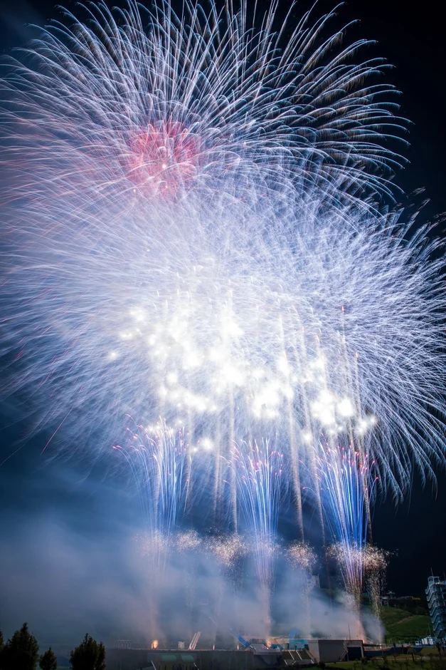 夜空に咲き乱れる壮大な花火大会の様子。青、白、赤を基調とした色とりどりの光が夜空を埋め尽くし、下からは打ち上げ花火が勢いよく立ち上り、煙が幻想的な雰囲気を醸し出しています。