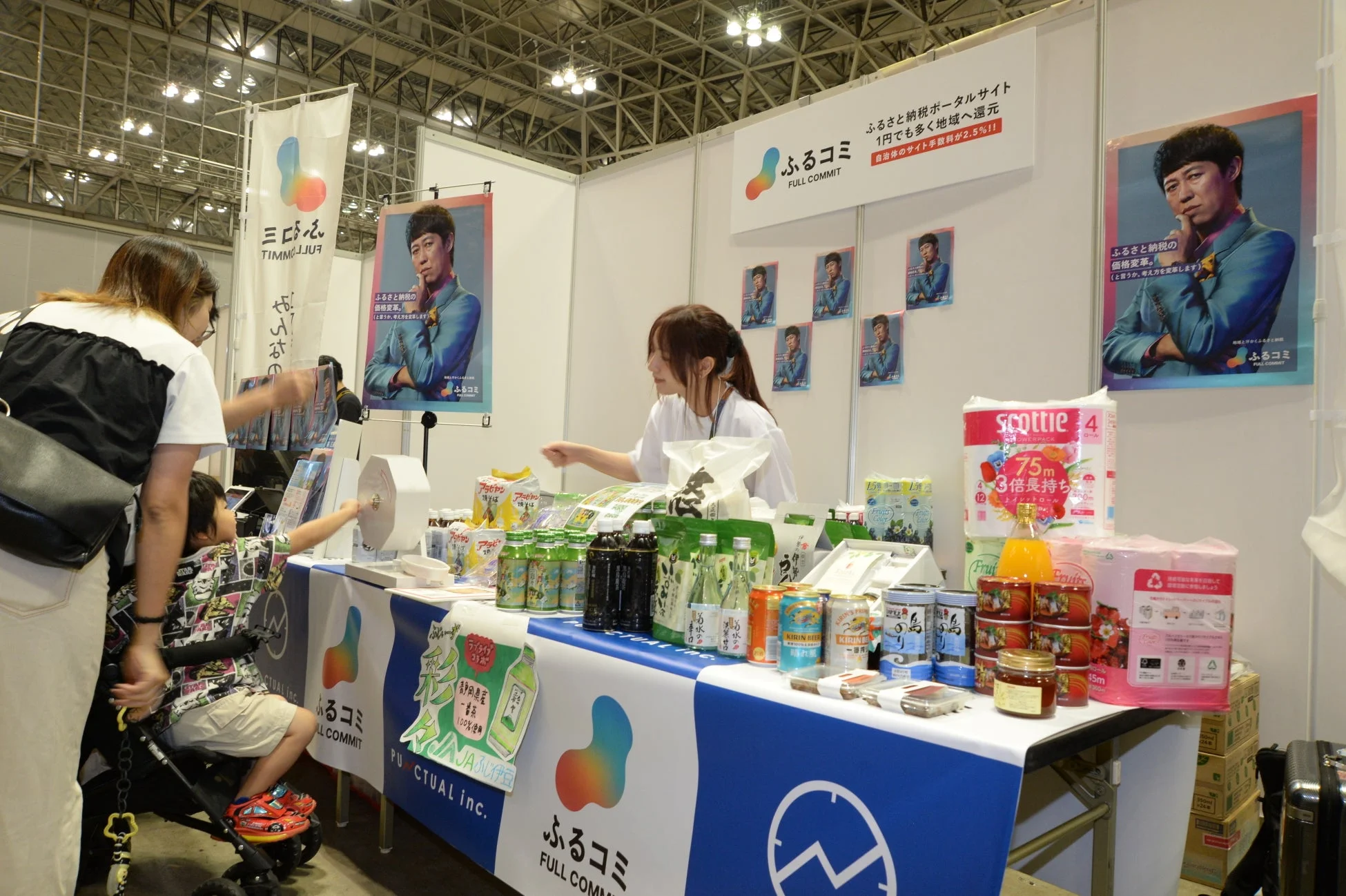 ふるさと納税ポータルサイト「ふるコミ」の展示ブースで、スタッフが来場者に対応している。ブースには返礼品らしき商品が多数並び、女性と子供が興味深そうに見ている。男性タレントのポスターも目立つ、活気あるイベントの様子。