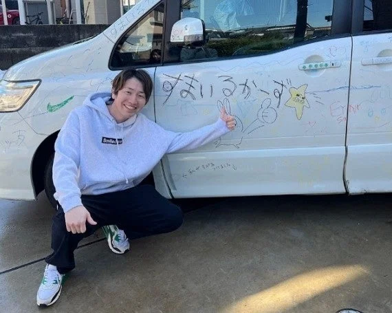白いミニバンに子供たちの描いた落書きやメッセージがびっしり書かれており、その車の前で笑顔の男性がサムズアップポーズをしている。